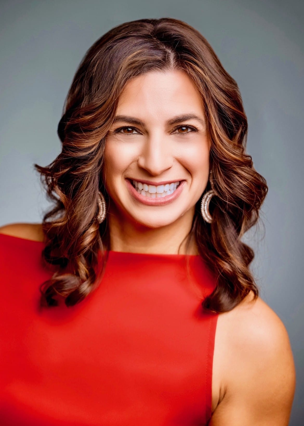 Smiling woman with wavy brown hair in a red sleeveless dress and hoop earrings, facing the camera against a neutral backdrop.