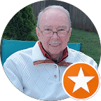 Smiling older man with glasses wearing a light blue jacket, seated outdoors on a blue bench, orange star badge in bottom-right corner.
